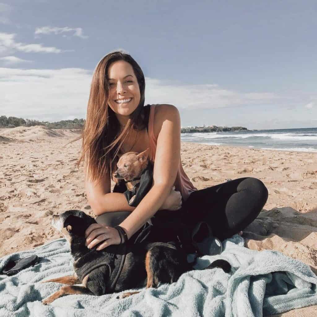 Image of a young lady relaxing on Culburra Beach South Coast NSW. She is cuddling one small dog in her arms and patting another small dog on the beach.
