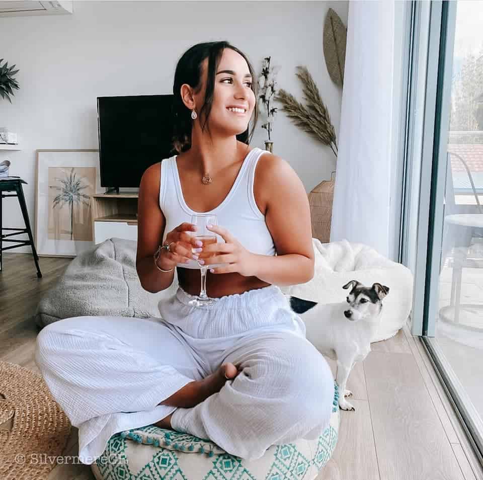 A young lady wearing all white siting in a yoga position on a cushion in the living room of Culburra Beach South Coast NSW. She is in the living room with her dog enjoying the views of the ocean, beach and lake.