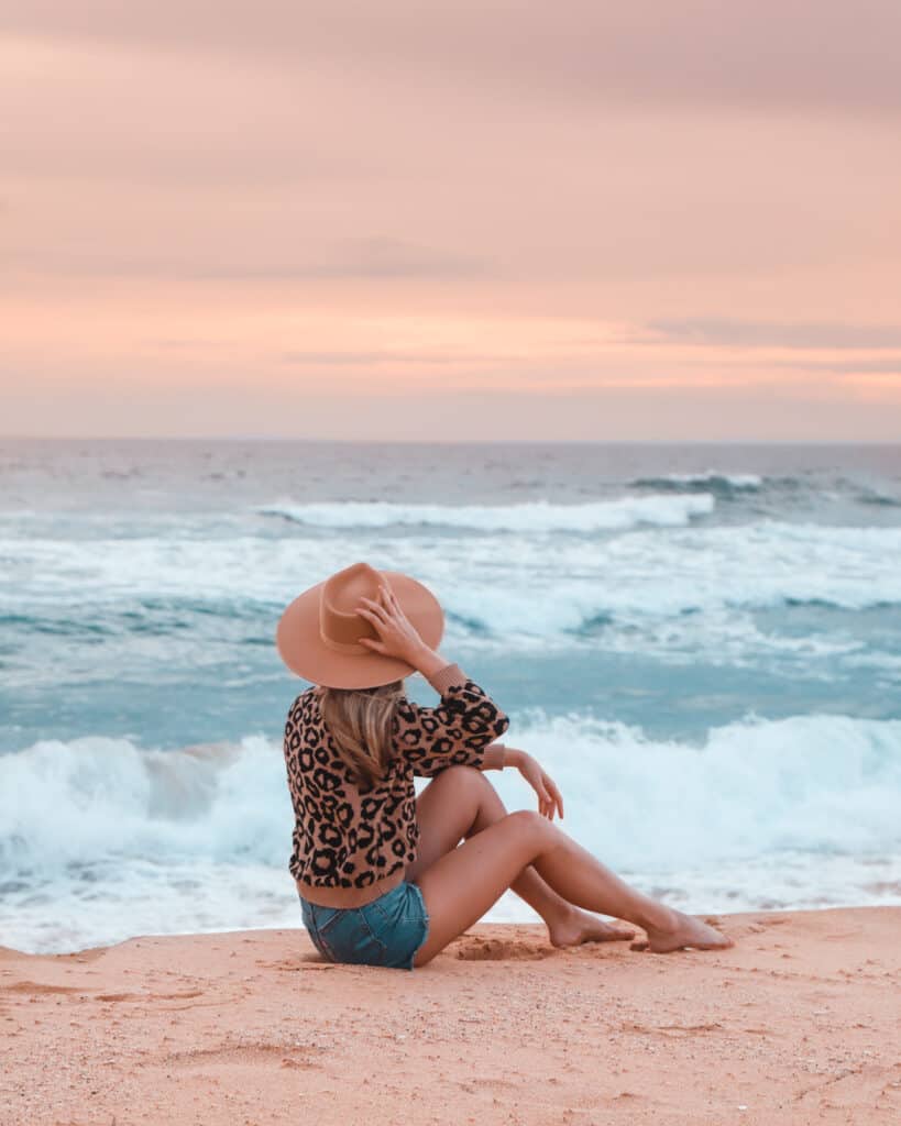A young lady relaxing on Culburra Beach on the South Coast NSW, holding her fedora hat, watching the waves roll in. The sun is setting in the background and has a beautiful pink to it.