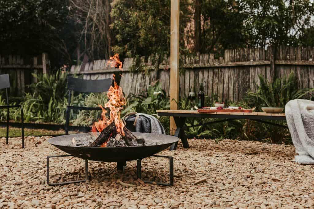 Fire burning in the fire pit in the fire pit area in the rear yard of Silvermere Coastal Retreat Culburra Beach South Coast NSW. The fire pit is surrounded by chairs and a bench. On the bench is some snacks, red wine and a blanket.