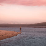 A young lady standing and enjoying Lake Wollumboola at Culburra Beach South Coast NSW. The lake is a bit rough with the sun setting in the background causing a pink hue.