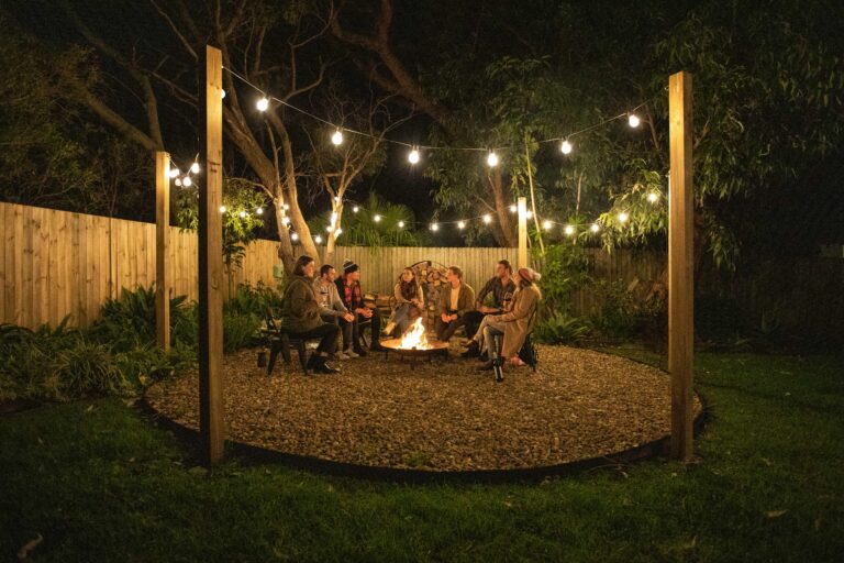 Book now - Fire pit area located in the rear of Silvermere Coastal Retreat located in Culburra Beach on South Coast NSW. The fire pit has a bunch of friends sitting around a firepit on pebbles. Festoon lights light up the area which is located under a beautiful canopy of local trees. The beach can be heard from the fire pit.