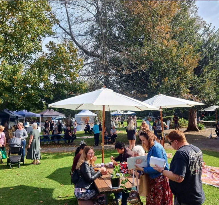 Summertime fun markets are located outside of Jindy Andy Mill in Pyree on South Coast NSW. People are sitting around on wooden seats and chairs enjoying the sun and gardens.