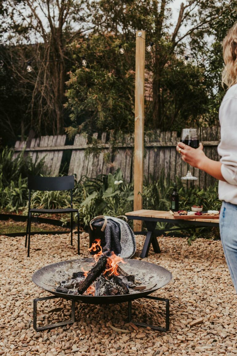 Women drinking red wine by the fire pit in the rear yard of Silvermere Coastal Retreat Culburra Beach South Coast NSW. Next to the fire is a bench seat with snacks, more wine and a blanket.