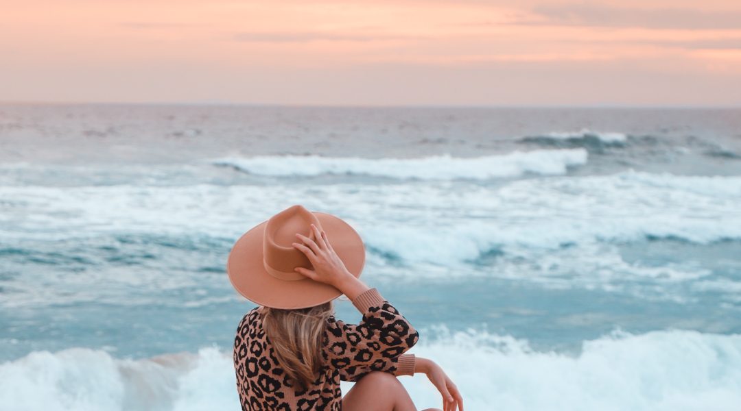A young lady relaxing on Culburra Beach on the South Coast NSW, holding her fedora hat, watching the waves roll in. The sun is setting in the background and has a beautiful pink to it.